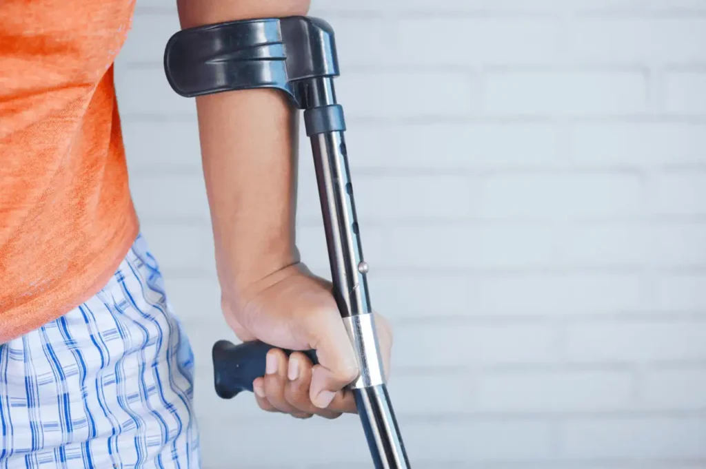 Person using a forearm crutch during post-surgery recovery, wearing casual clothes and standing against a white brick wall — representing active rehab support in Vancouver.
