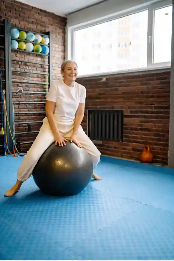 A smiling older woman sits astride a stability ball on a blue mat in a brick‑walled gym—ideal for seniors’ personal training and active rehab near me in Vancouver & Kitsilano, combining personalised kinesiology with fall prevention.